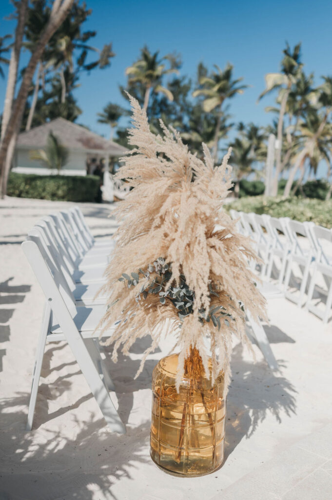 Large glass vase full of pampas grass beside row of white folding chairs at beach wedding ceremony | Lauren E. Bliss Destination Wedding Photography