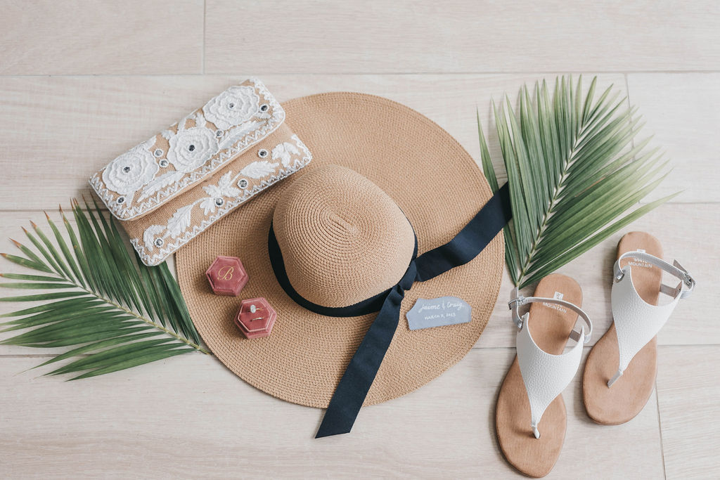 Sunhat, sandals, and white patterned clutch purse laying next to palm leaves