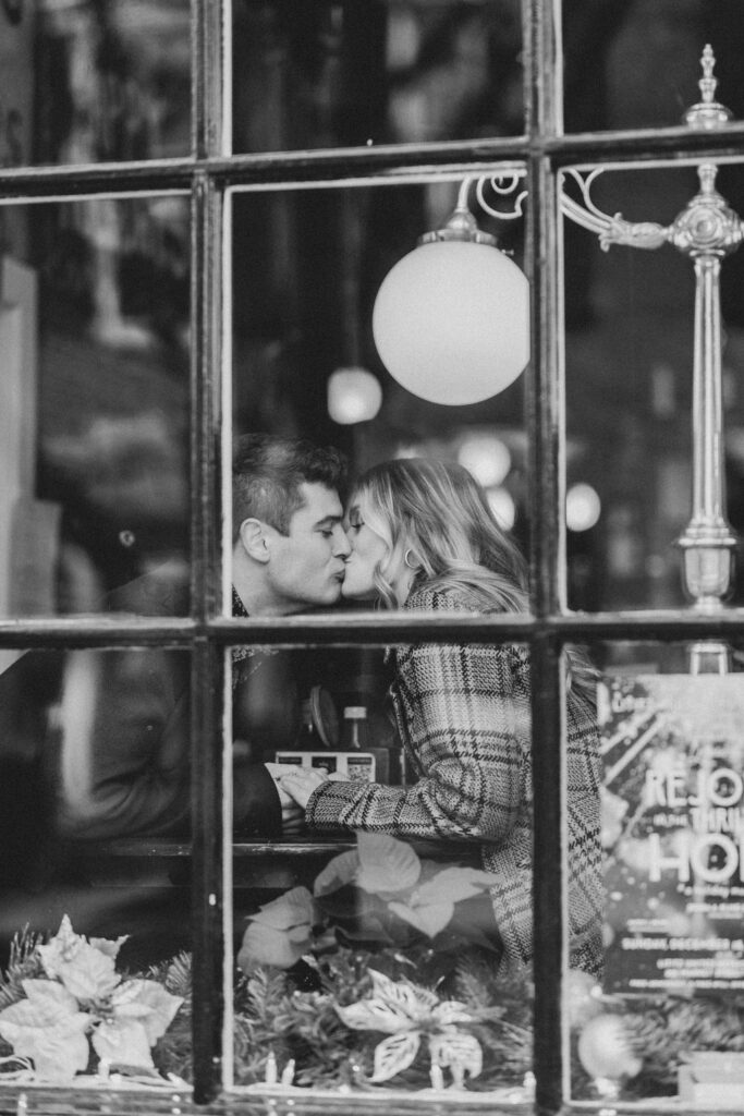 Black and white photo taken through a window of couple kissing at a table inside of Bulls Head Public House in Lititz | Dowtown Lititz Engagemnent Photo Session by Lauren E. Bliss Photography