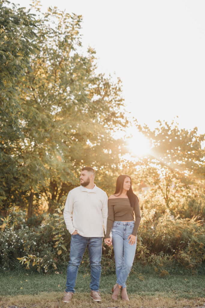 Couple holds hands and looks in opposite directions in front of woodsy park sunset | Overlook Park Engagement Photo Session by Lauren E. Bliss Photography