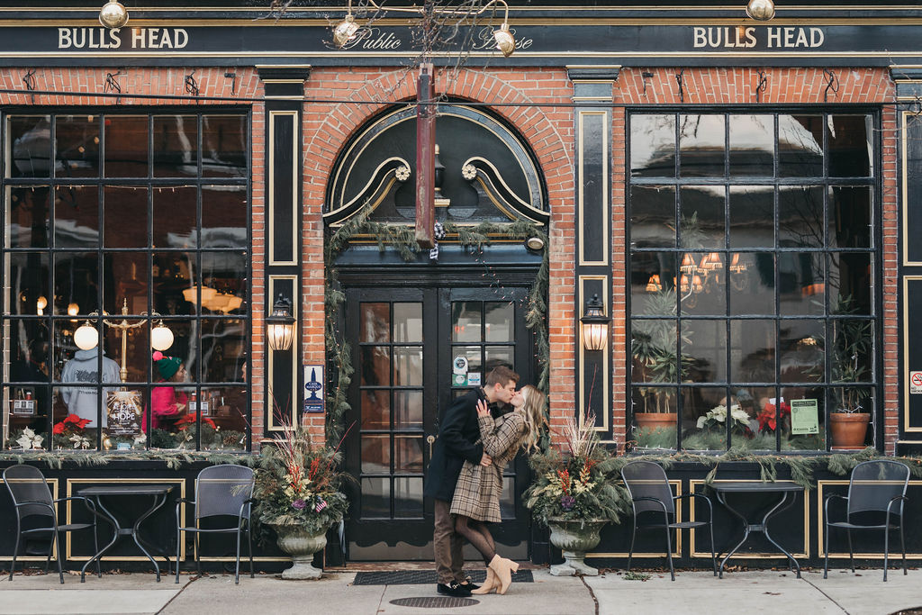 Couple kissing in front of Bulls Head Public House in Lititz decorated with Christmas decor | Dowtown Lititz Engagemnent Photo Session by Lauren E. Bliss Photography