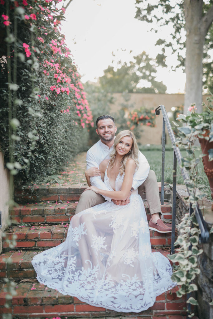 Couple sits on brick stairway and smiles at camera in front of live pink floral hedge in background | Conestoga House and Gardens Engagemnent Photo Session by Lauren E. Bliss Photography