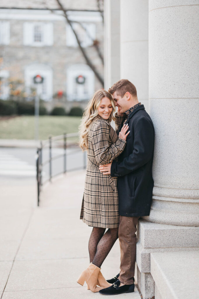 Couple standing in front of large stone columns with stone building decorated with Christmas wreaths in the background |Downtown Lititz Engagement Photo Session by Lauren E. Bliss Photography