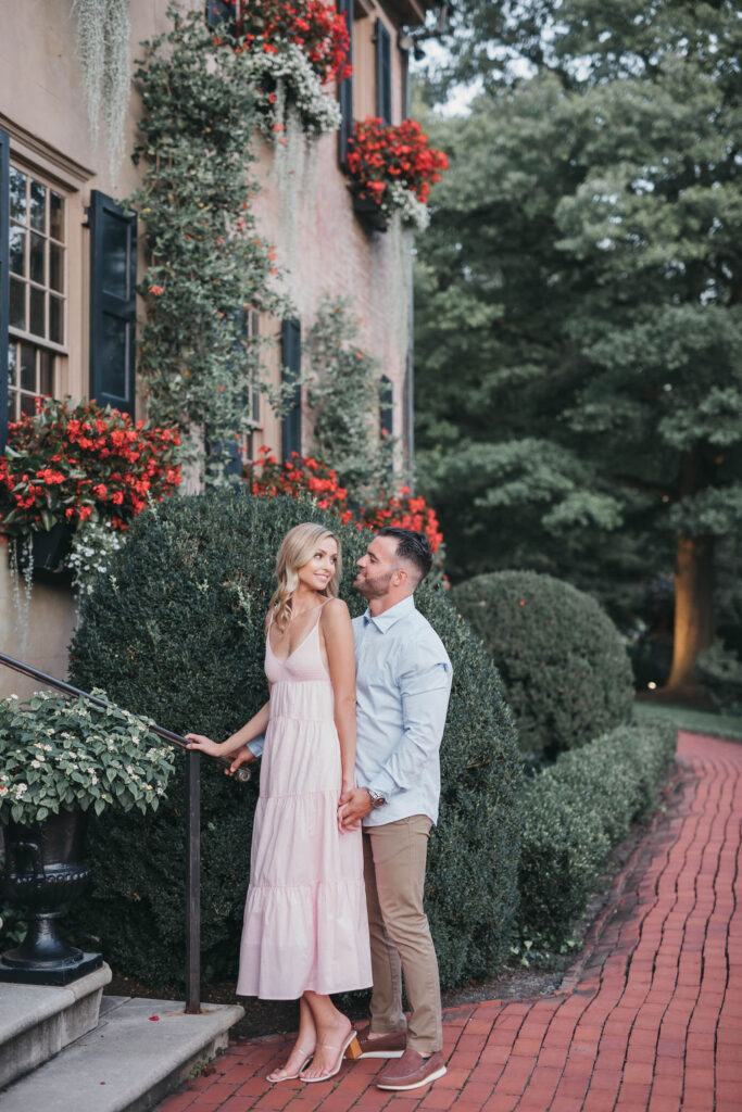 Couple stands on brick pathway and smiles at each other with red flower window boxes in background | Conestoga House and Gardens Engagemnent Photo Session by Lauren E. Bliss Photography