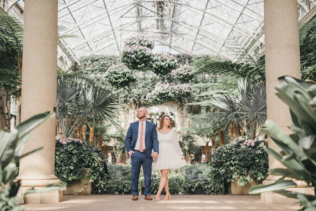 Man and woman stand hand in hand facing opposite directions in front of landscaped florals inside Longwood Gardens greenhouse display