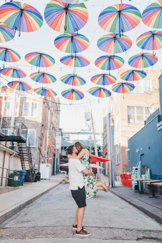 Man holds woman up as they kiss under hanging rainbow umbrellas in a city street | Downtown Manayunk Engagement Session by Lauren E. Bliss Photography