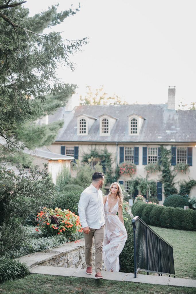 Man leads woman up a stone staircase with historic Lancaster County mansion in the background | Conestoga House and Gardens Engagemnent Photo Session by Lauren E. Bliss Photography