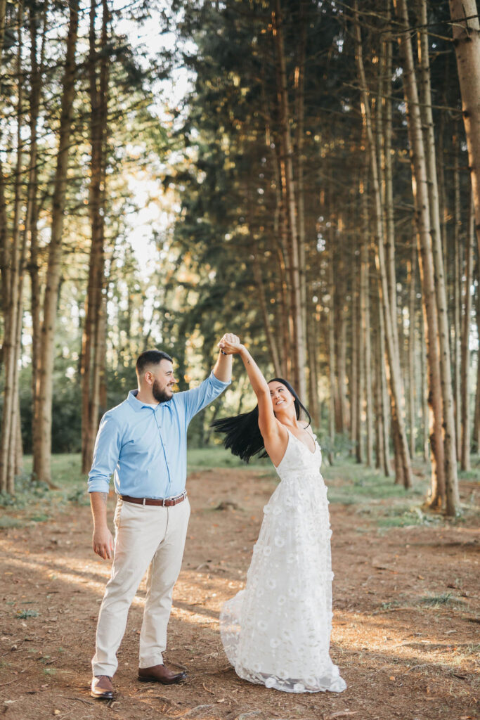 Man twirls fiance in a white floor length floral dress under a canopy of tall trees | Overlook Park Engagement Photo Session by Lauren E. Bliss Photography