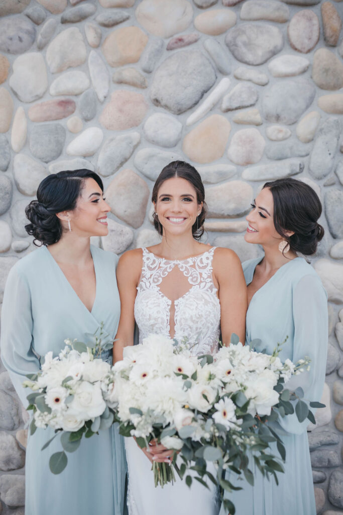 A bride wearing white in between two bridesmaids wearing light blue long sleeved dresses in front of a gray stone wall | Lauren E. Bliss Photography Luxury PA Wedding Photographer