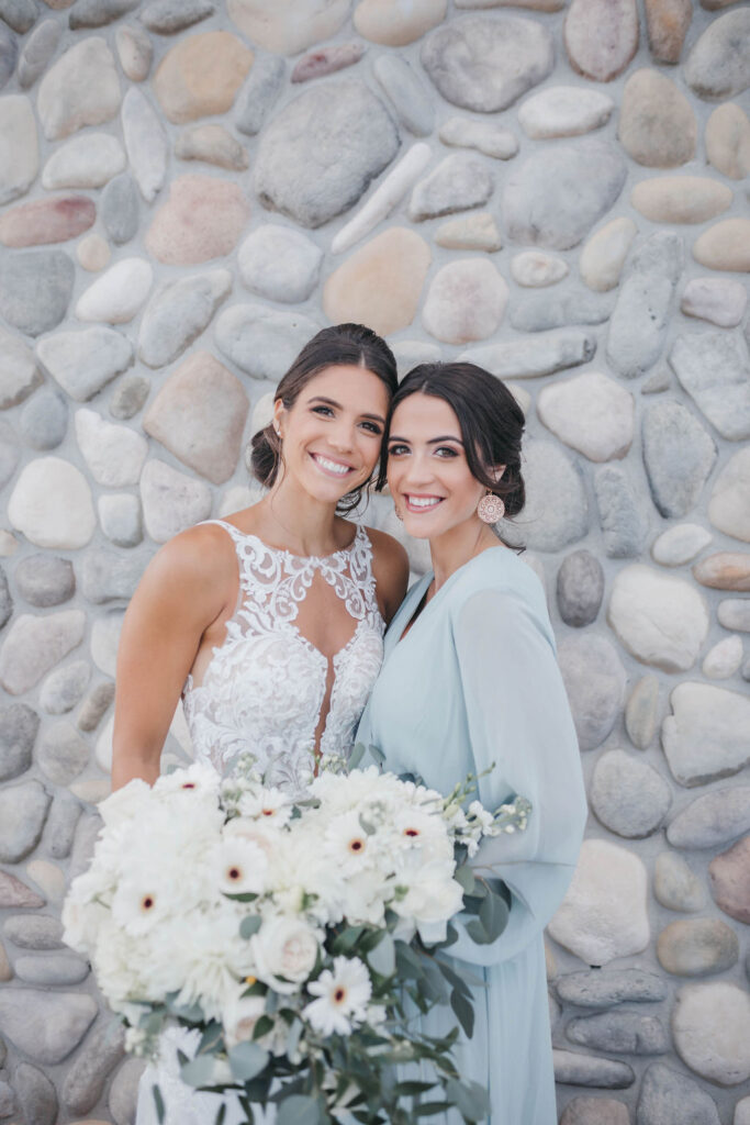A bride in white and her bridesmaid in a long sleeved light blue dress holding hands and smiling at the camera in front of a gray stone wall | Lauren E. Bliss Photography Luxury PA Wedding Photographer