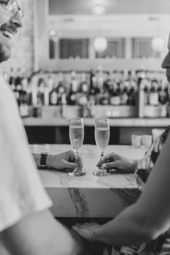 Two people sit at a white marble bar facing each other with two full champagne glasses in between them | Downtown Manayunk Engagement Session by Lauren E. Bliss Photography