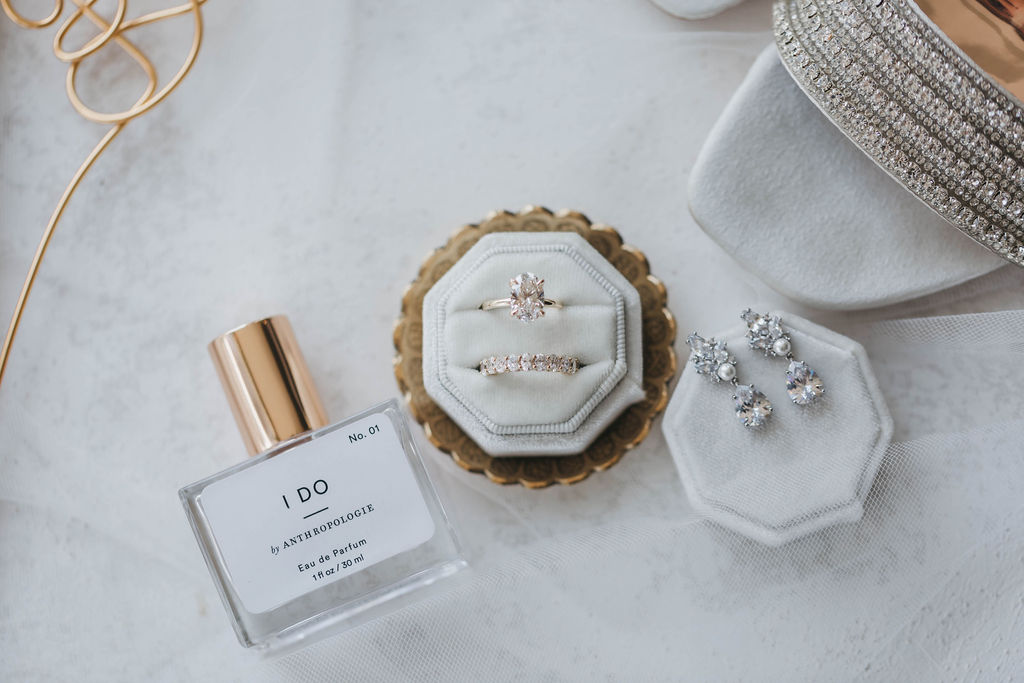 I Do perfume from Anthropologie lays beside a beige velvet ring box holding a gold oval cut diamond engagement ring and diamond wedding band