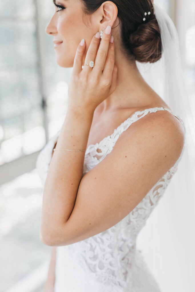 Bride smiling and looking off into the distance while brushing her cheek with her left hand and ring finger