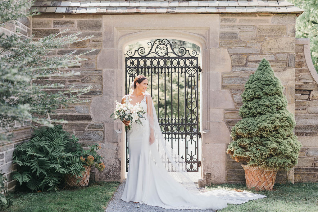 Bride holding white and green floral bouquet in front of wrought iron gateway at Greystone Hall venue in West Chester PA | Lauren E. Bliss Photography
