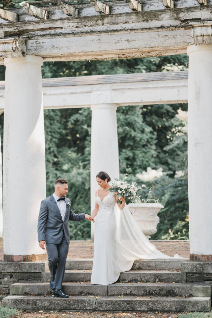 Groom leading bride down a flight of stone steps underneath a white column pergola at Greystone Hall wedding venue in PA