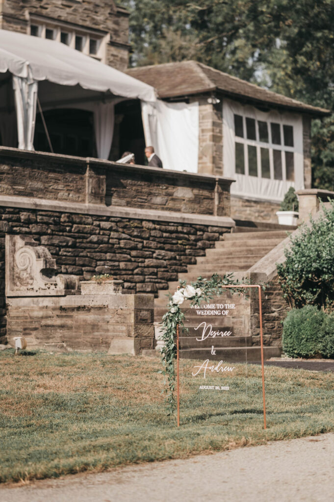 Acrylic wedding welcome sign sits on lawn in front of a flight of stone steps