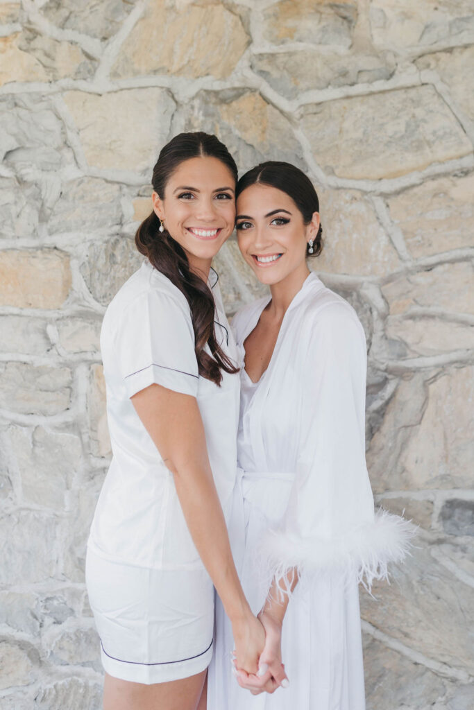 two girls wearing white holding hands and smiling at the camera in front of a gray stone wall | Lauren E. Bliss Photography Luxury PA Wedding Photographer