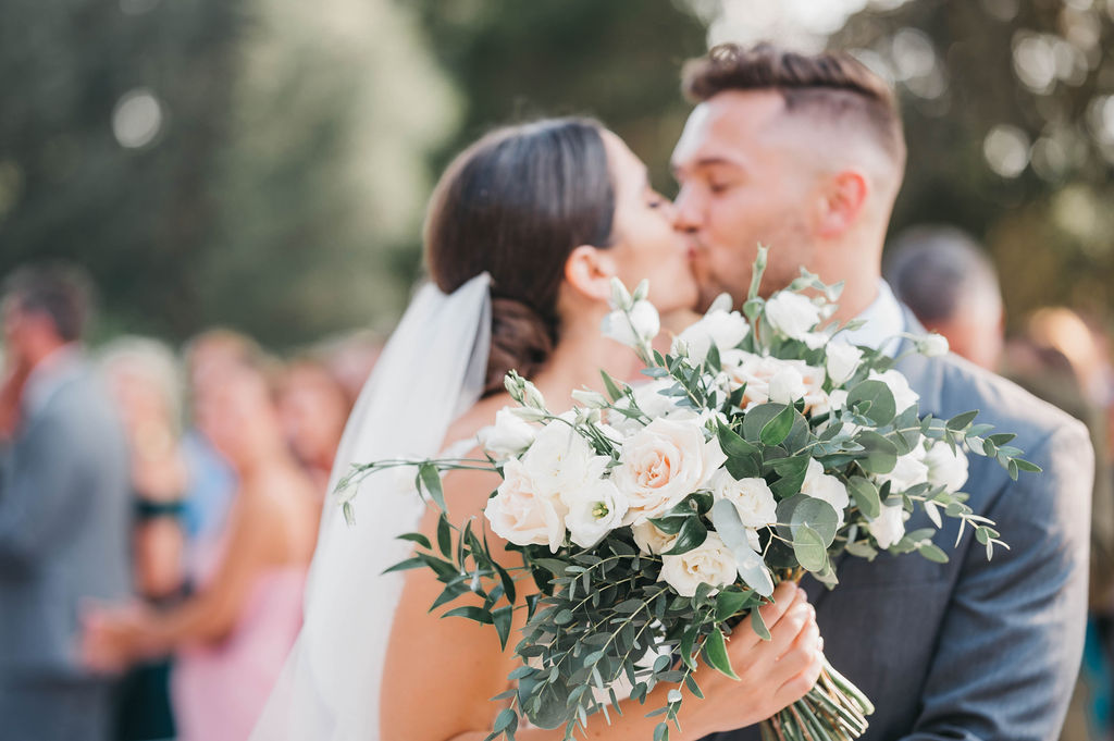 Bride and groom are kissing and out of focus while the focal point is on the brides white floral and greenery bouquet