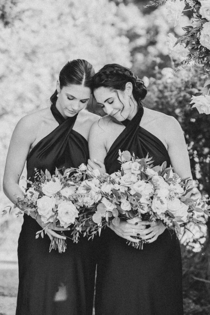 Two matrons of honor hold bouquets and rest their heads together with their eyes closed