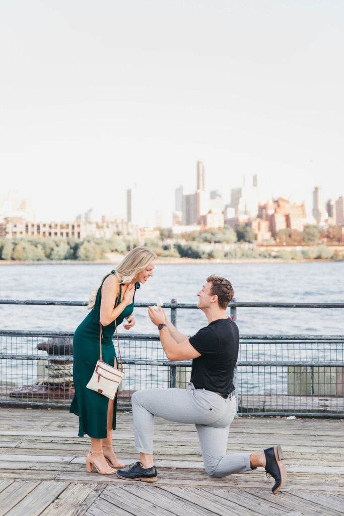 Man on one knee proposes to girlfriend on a pier overlooking the Brooklyn Bridge on  the East River in NYC