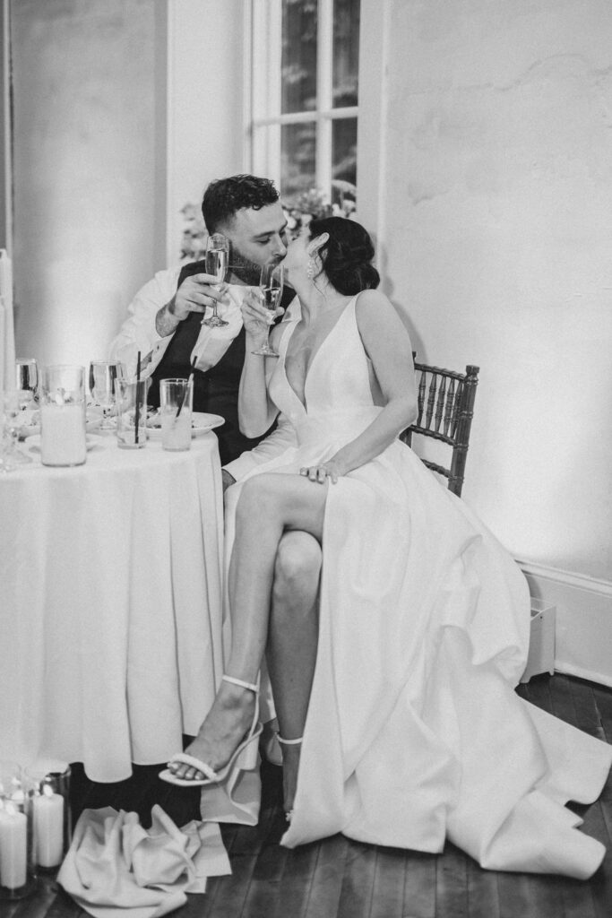 Black and white photo of bride and groom sitting at sweetheart table and kissing at wedding reception