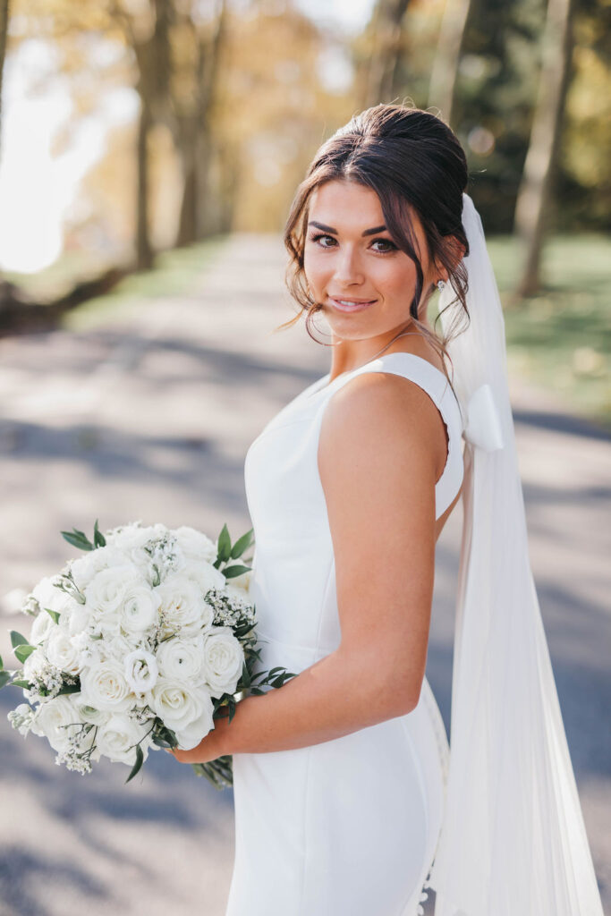 Bride in white dress looks over her shoulder while holding a round bouquet of white roses and baby's breath