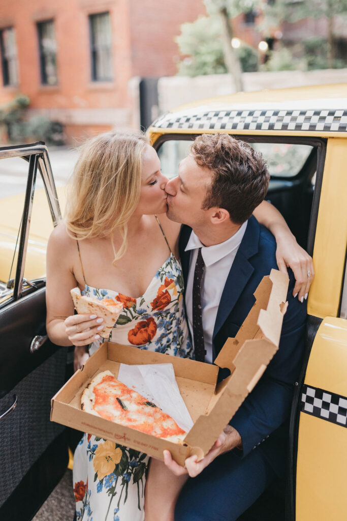 Man and woman sit in a yellow taxi holding a box and slices of NYC pizza as they kiss