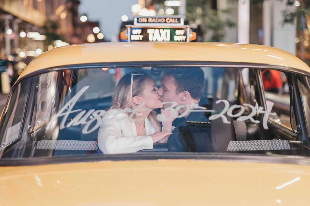Man and woman kiss in the backseat of a yellow NYC taxi with the date August 30, 2024 written on the rear windshield 