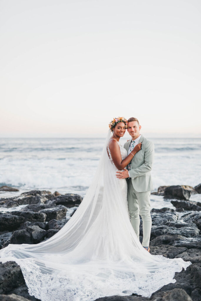 Bride and groom stand on black rocks at the edge of the water during their Hawaii destination wedding
