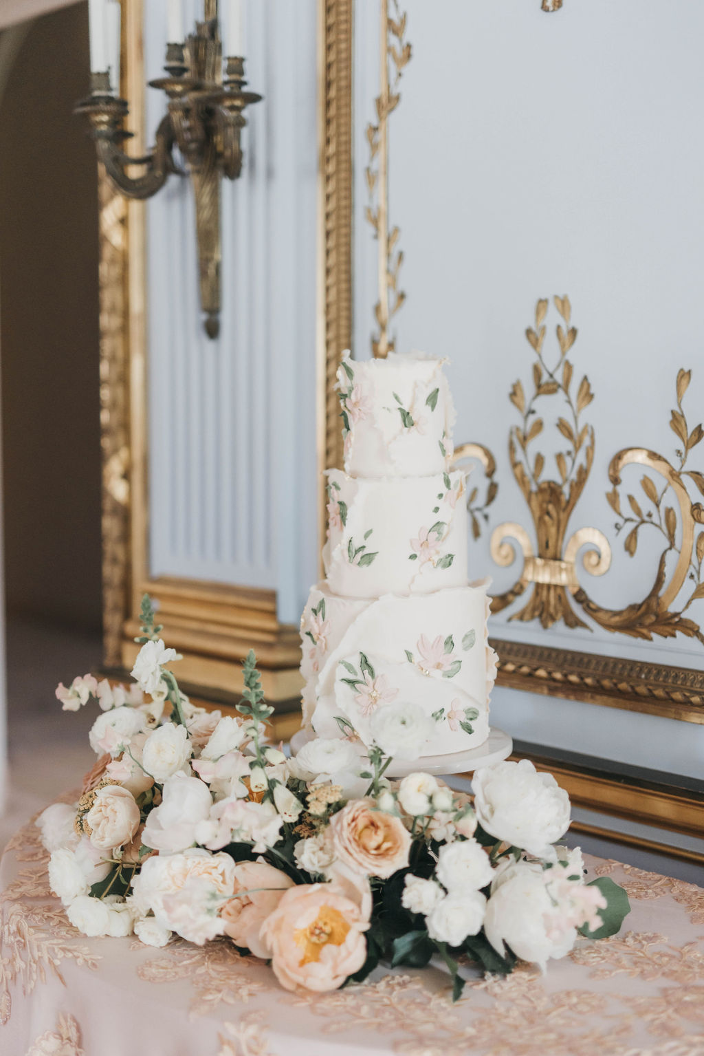 Elegant three tiered white wedding cake features sugar flowers and intricate piping design and is surrounded by fresh flowers on a small side table at Elkins Estate in Philadelphia