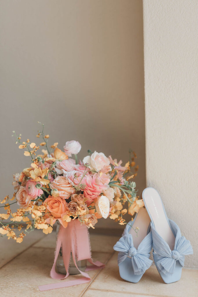 Pastel pink and orange hand tied wedding bouquet sits in a vase beside light blue Loeffler Randall heels