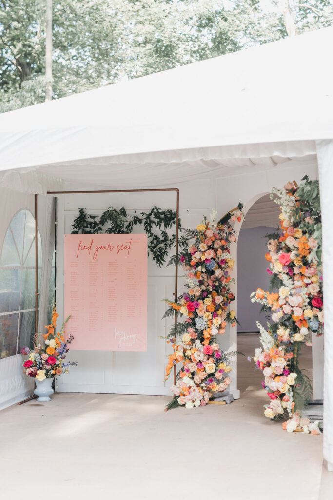 Colorful pink orange and yellow floral arch next to pink seating chart in front of reception entryway | Colorful wedding photography by Lauren E. Bliss Photography