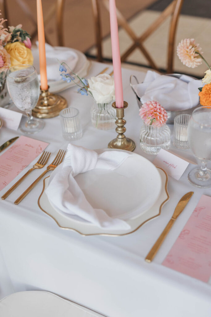 Brightly colored pink and orange tapered candles in brass candleholders on a white linen covered banquet table