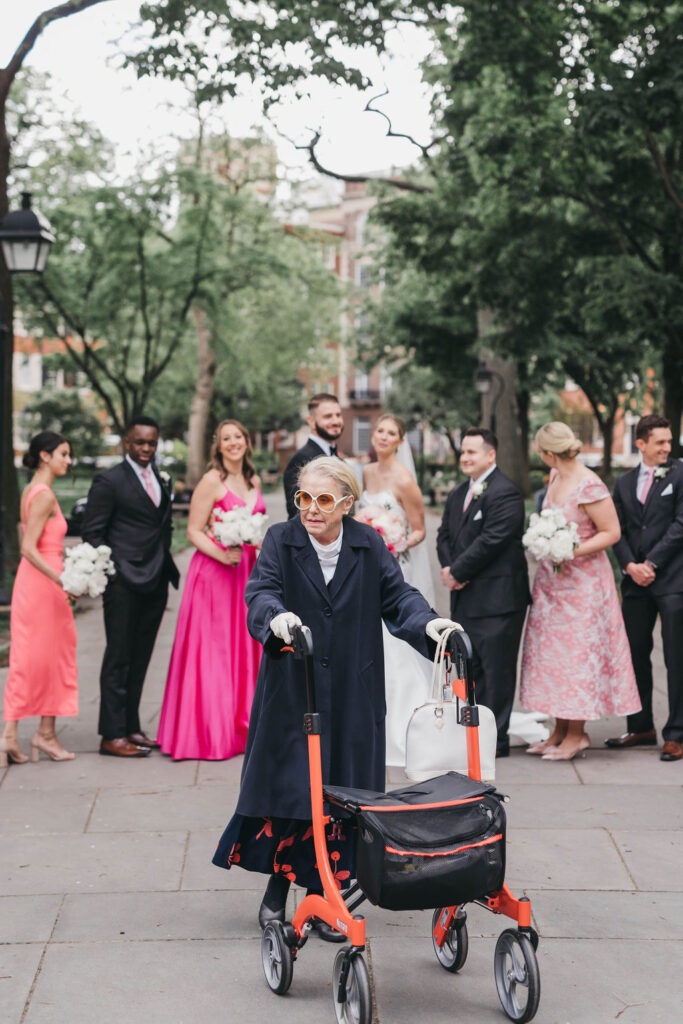 Old woman steers a red rolling walker in front of a bridal party on stone sidewalk