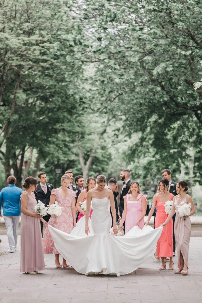 Bridesmaids wearing dresses in different shades of pink surround the bride and lift the train of her mermaid style wedding dress