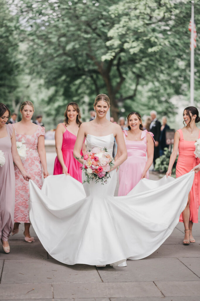 Bridesmaids wearing dresses in different shades of pink surround the bride and lift the train of her mermaid style wedding dress