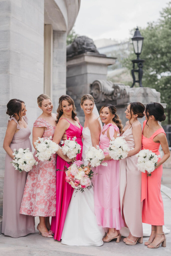 Three bridesmaids wearing dresses in different shades of pink hold white floral bouquets and stand on either side of a bride wearing a mermaid style wedding dress