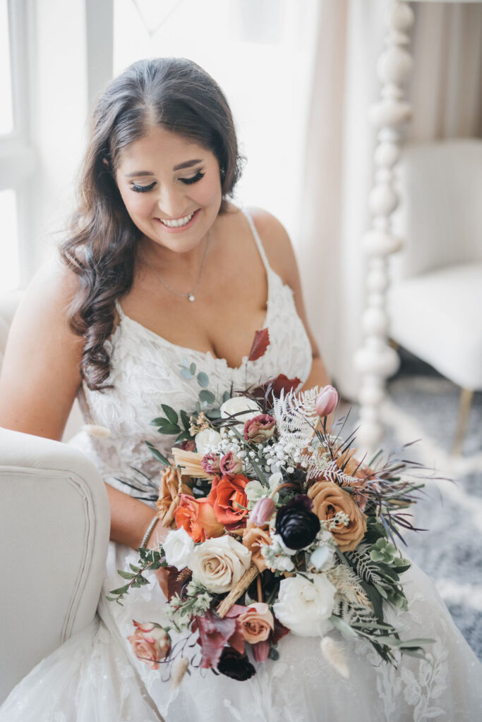 Bride sits and poses fall color wedding bouquet in lap while smiling and looking down