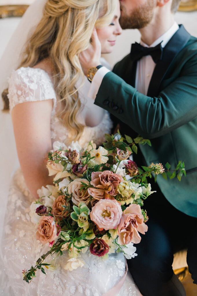 Groom in emerald green dinner jacket kisses bride as she looks down and holds a hand tied wedding bouquet of muted fall colors