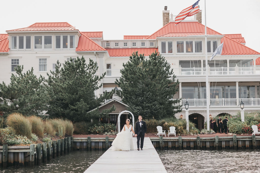 Bride and groom walk on a dock over the water in front of the Mallard Island Yacht Club wedding venue