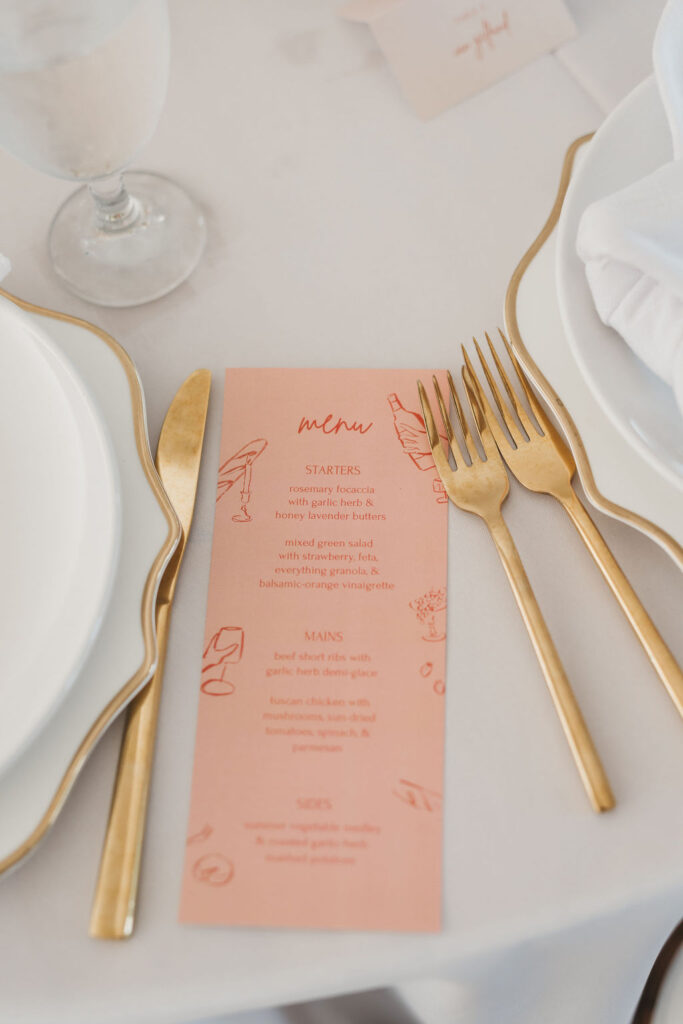 Peach colored wedding dinner menu sits on banquet table beside gold wedding flatware