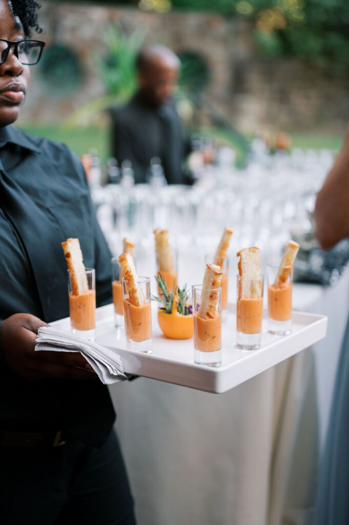 Member of caterer staff wearing all black carries a white tray of tomato soup shooters garnished with breadsticks at Winterthur Museum and Gardens