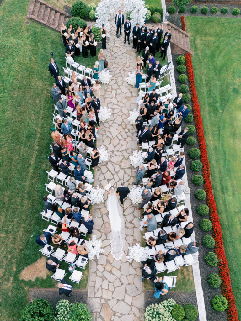 Aerial view of an outdoor wedding ceremony at Hamilton Farm Golf Club, showing white ceremony chairs, a stone aisle, lush floral arrangements, and guests gathered for a formal garden wedding.
