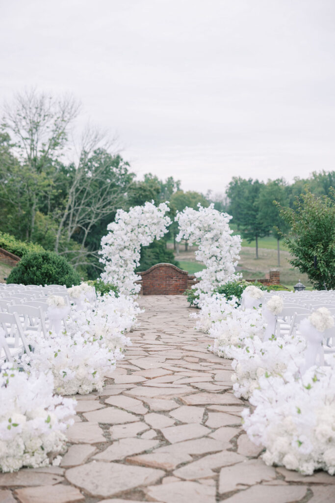 Stone ceremony aisle outdoors lined with white florals and white chairs leads to a large white floral arch at Hamilton Farm Golf Club