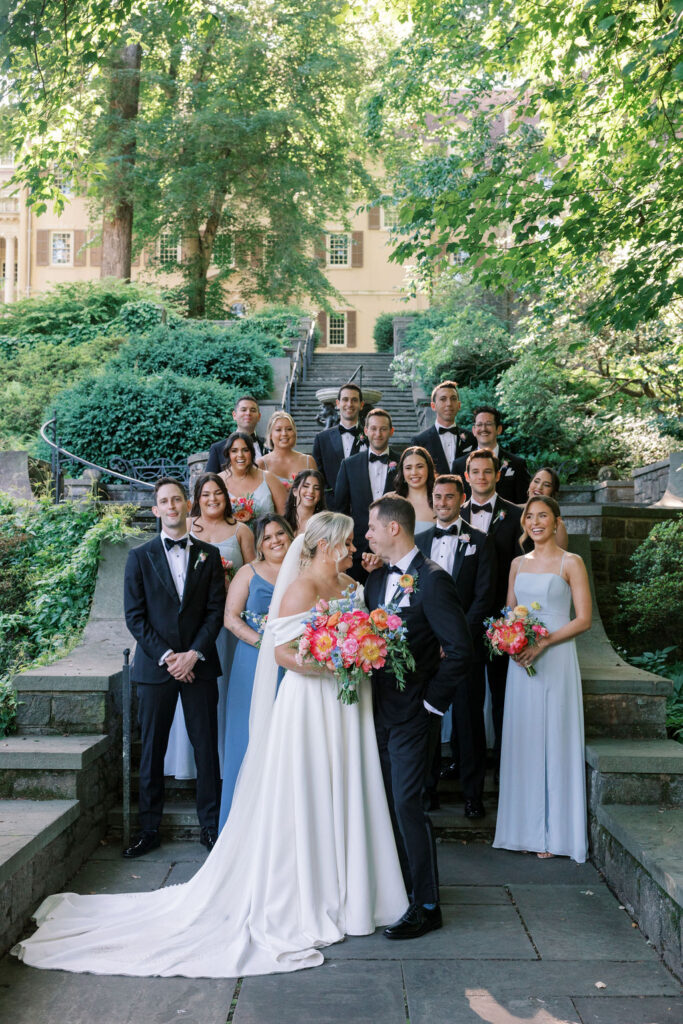 Bridal party wearing blue and black flanks a bride and groom on either side posing for wedding photos on the stone steps outside Winterthur Museum and Gardens