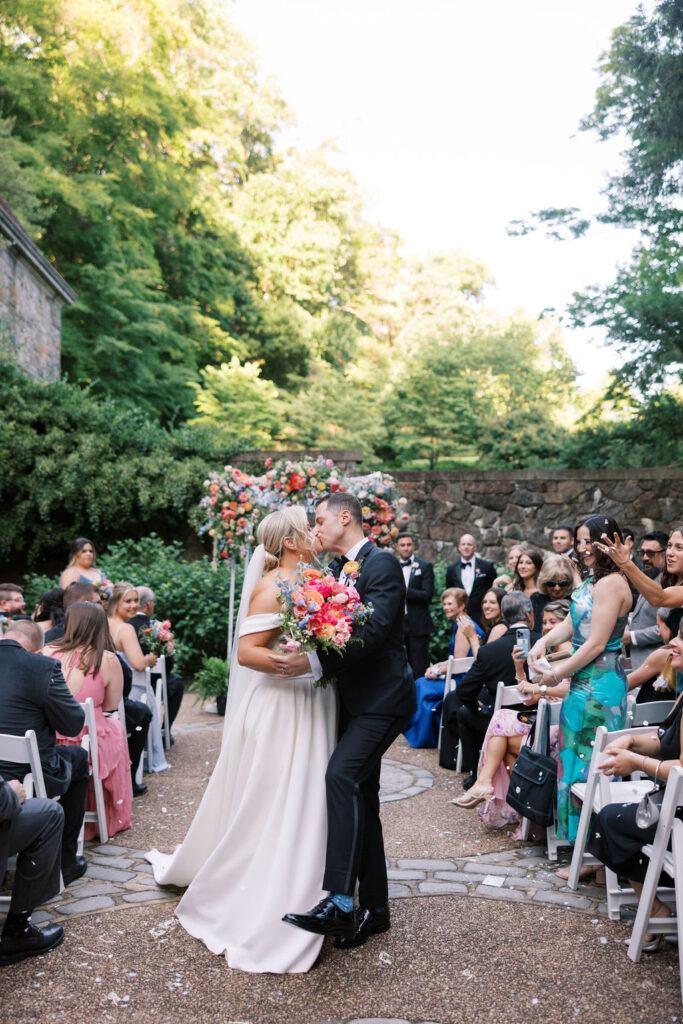 Bride and groom kiss in the middle of the outdoor ceremony aisle as wedding guests cheer and throw flower petals at Winterthur Museum and Gardens