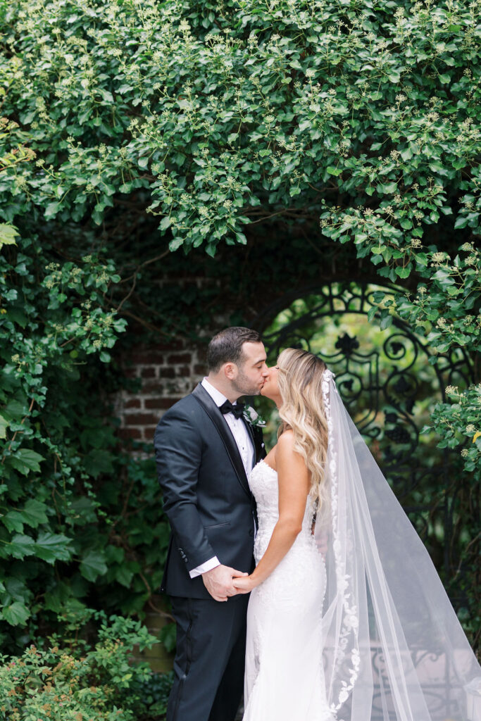 Bride and groom kiss under wall of trailing vines and wrought iron gate at Hamilton Farm Golf Club