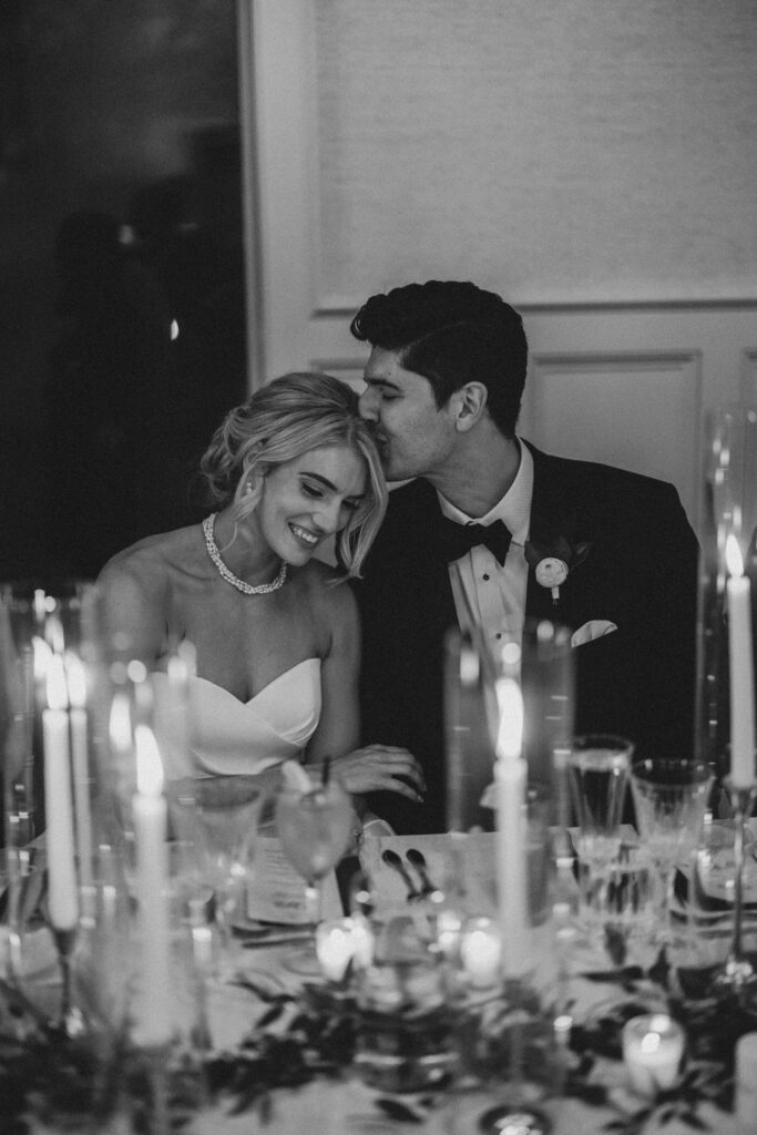 Groom kisses brides head while they sit at wedding reception dinner | Lauren Bliss Photography at Elkins Estate in Philadelphia