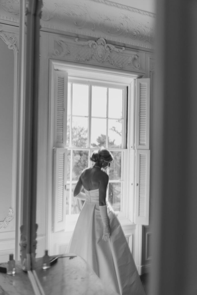 Bride wearing ballgown and gloves stands in front of window | Lauren Bliss Photography at Elkins Estate in Philadelphia