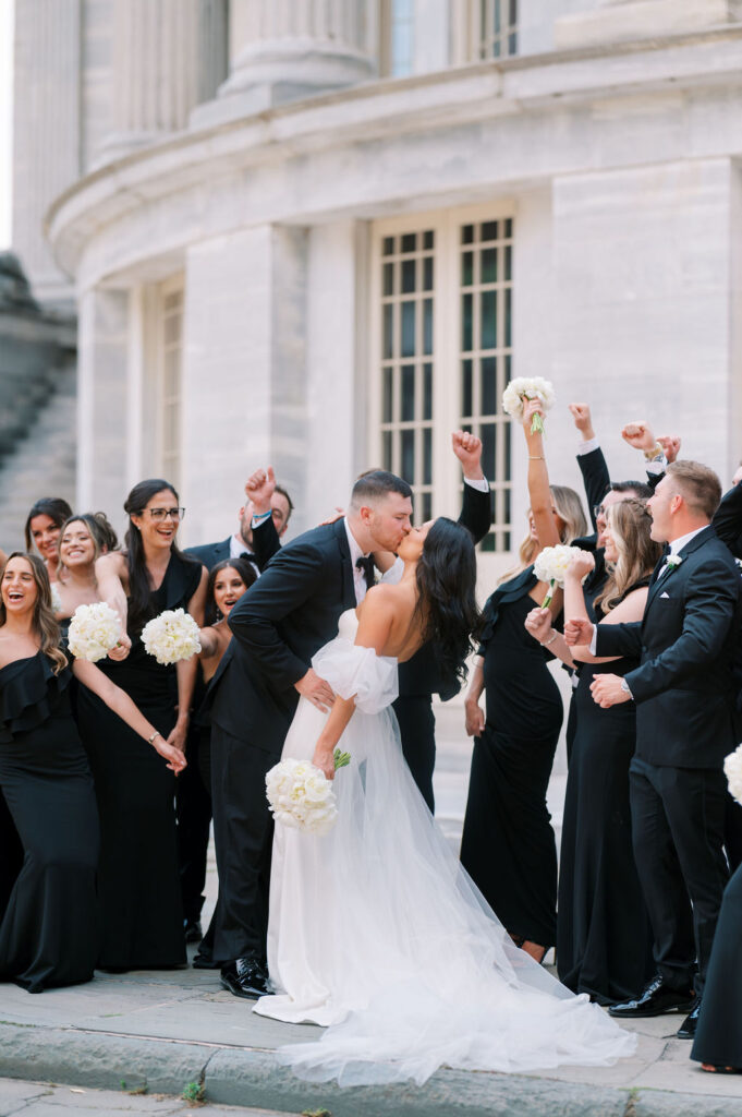 Bride and groom kiss in front of wedding party wearing all black | Wedding Photography by Lauren Bliss at the Cescaphe Ballroom Philadelphia